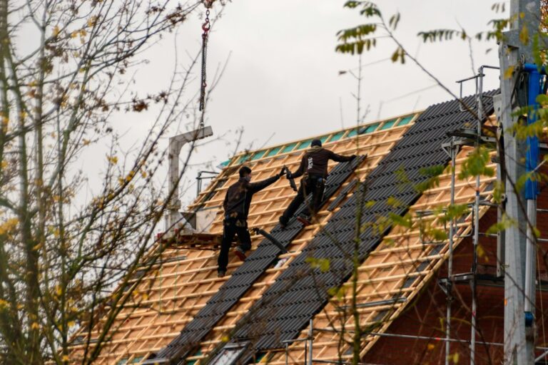 Workers remove old shingles from a house roof while protective tarps cover the ground below.