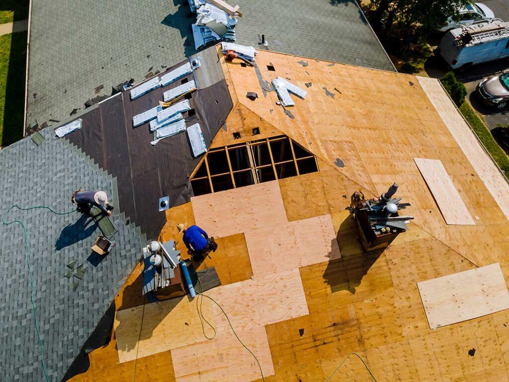 Workers protect a shingled roof with walk boards and secured covering beside a home renovation site.